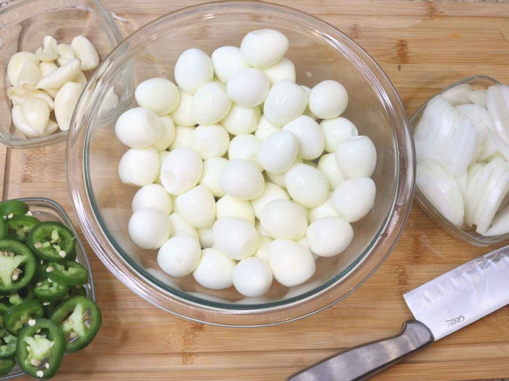 glass bowl of quail eggs, jalapenos, onions, garlic on wood cutting board with stainless knife beside them