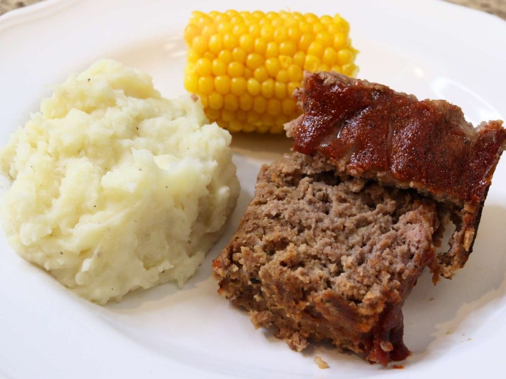 meatloaf with ketchup sauce, corn on the cob, and mashed potatoes on white plate