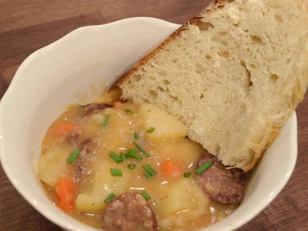 white bowl with sausage and potatoes in thick broth with piece of bread on the side