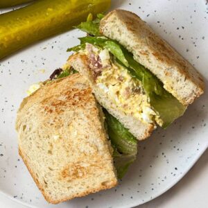 BLT egg salad sandwich cut in two sitting on a gray plate with pickles next to it